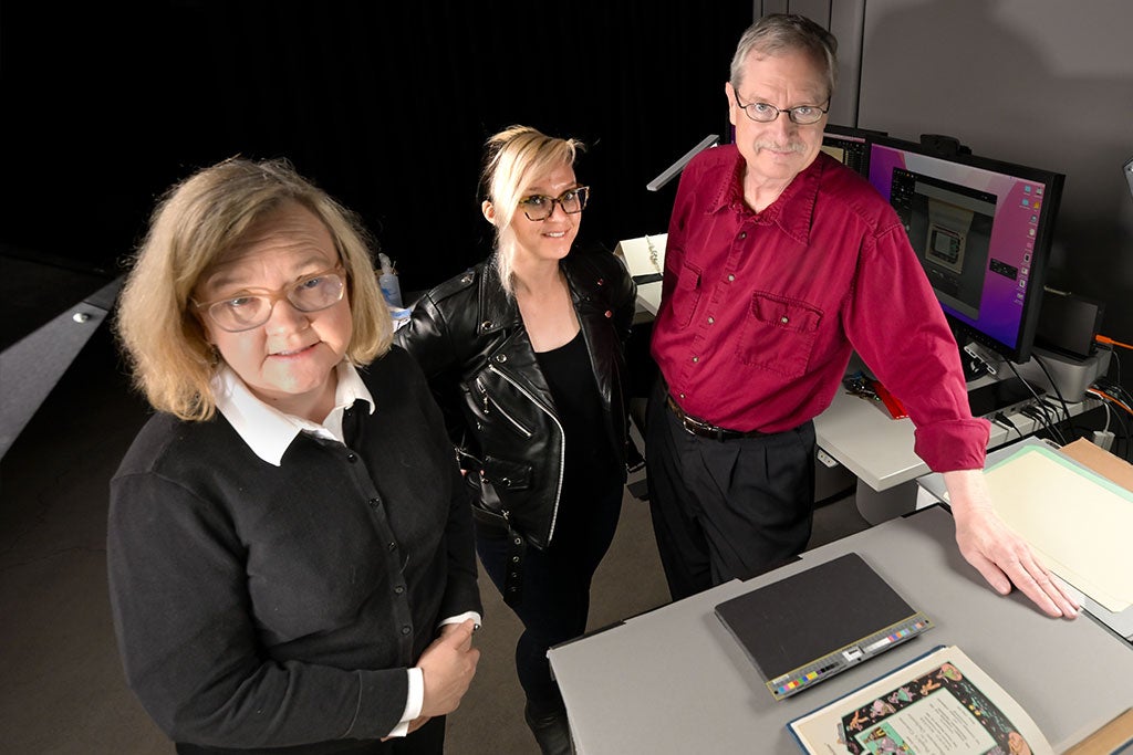 Three people in a tech room with computers, standing by a table with a book and papers.