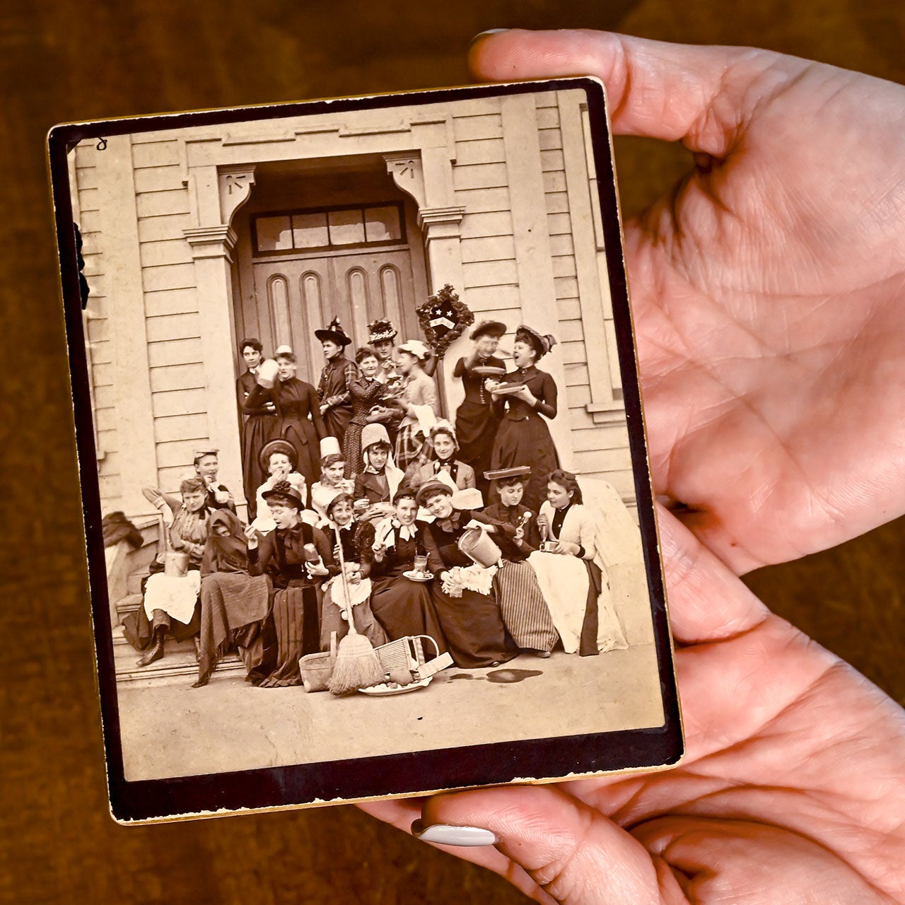 Hands hold an old sepia photo of a group of people in vintage clothing outside a building.