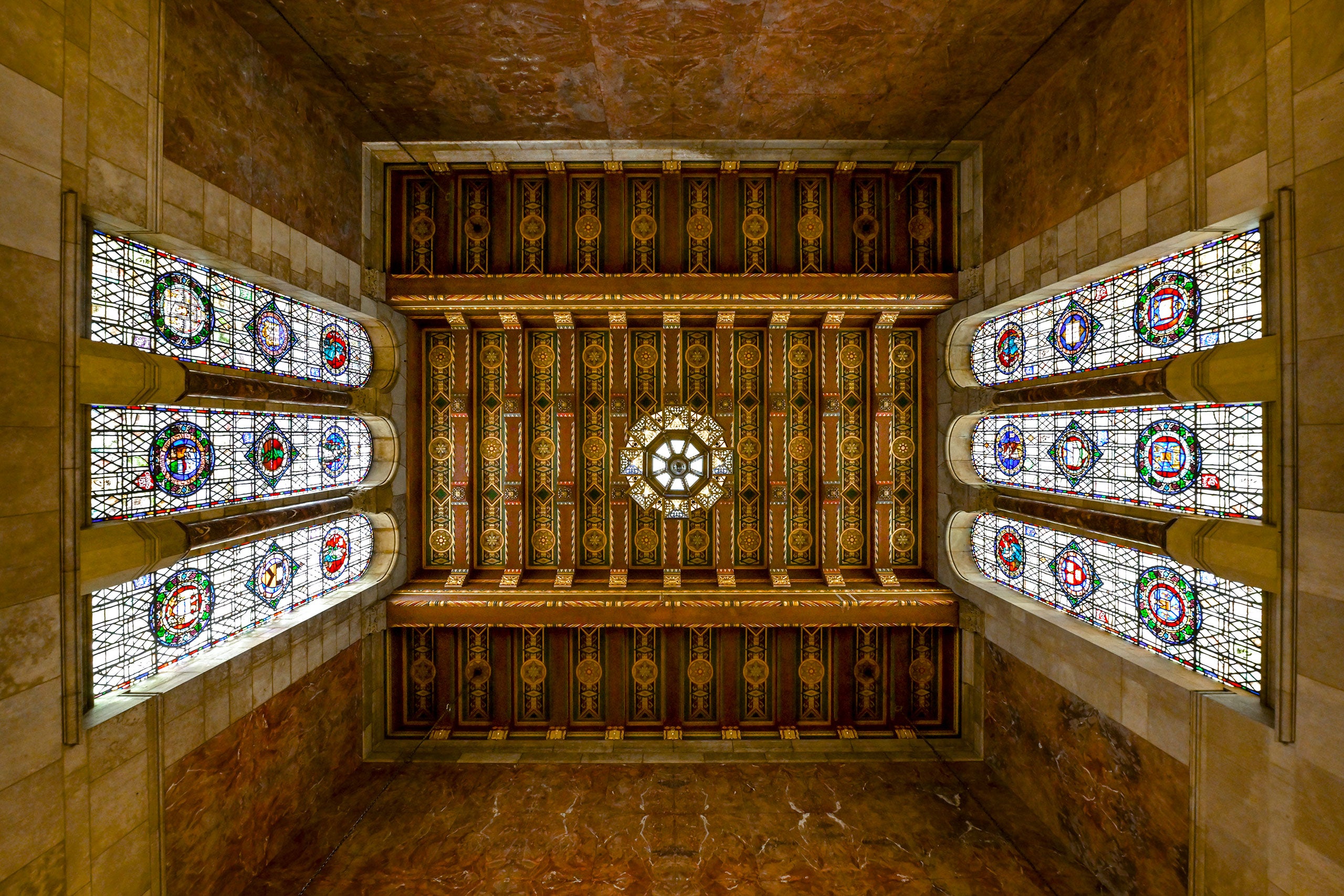 Ornate ceiling with stained glass windows and intricate patterns in a historic, stone-walled room.
