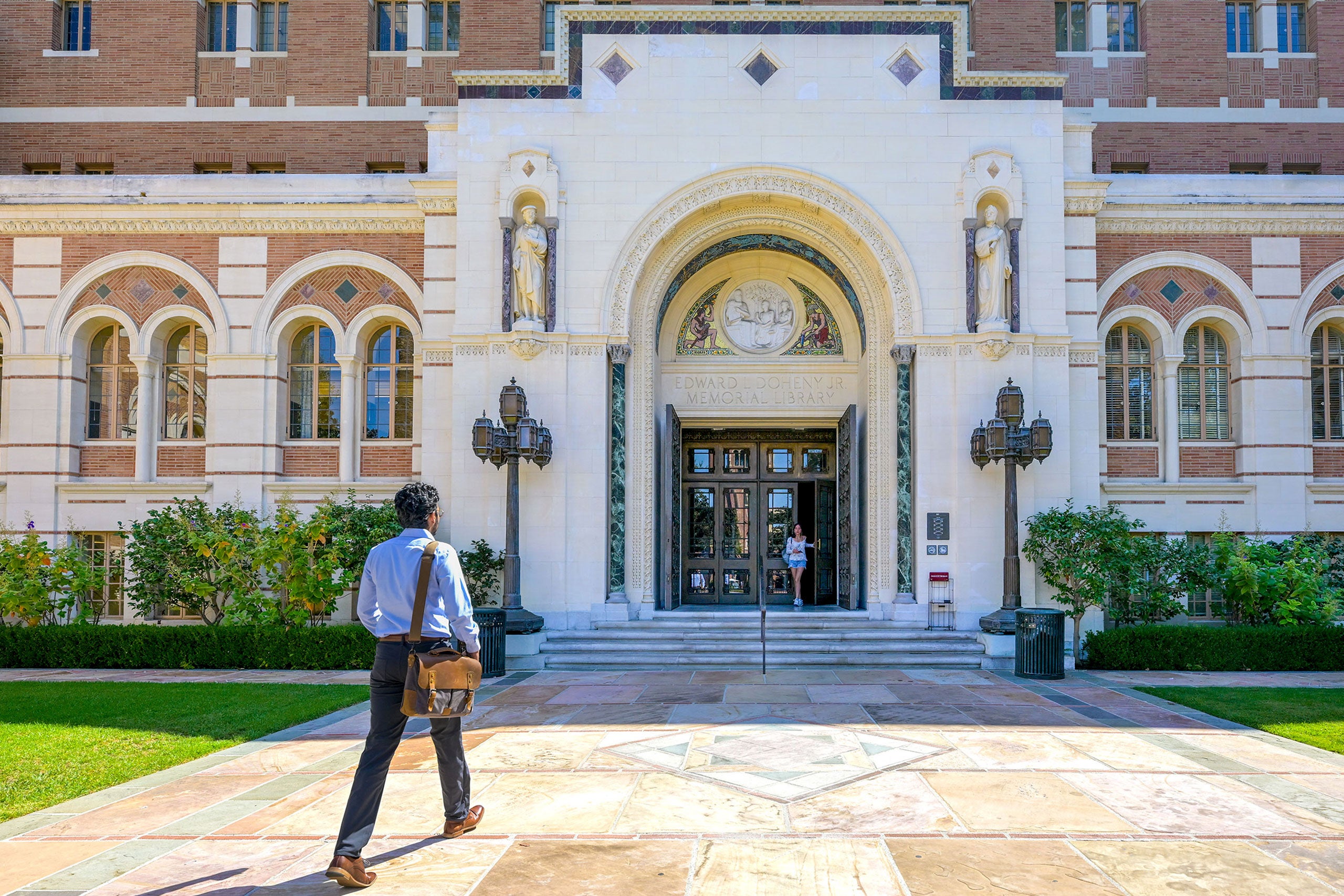 Person walks toward ornate library entrance with statues, arches, and greenery on a sunny day.