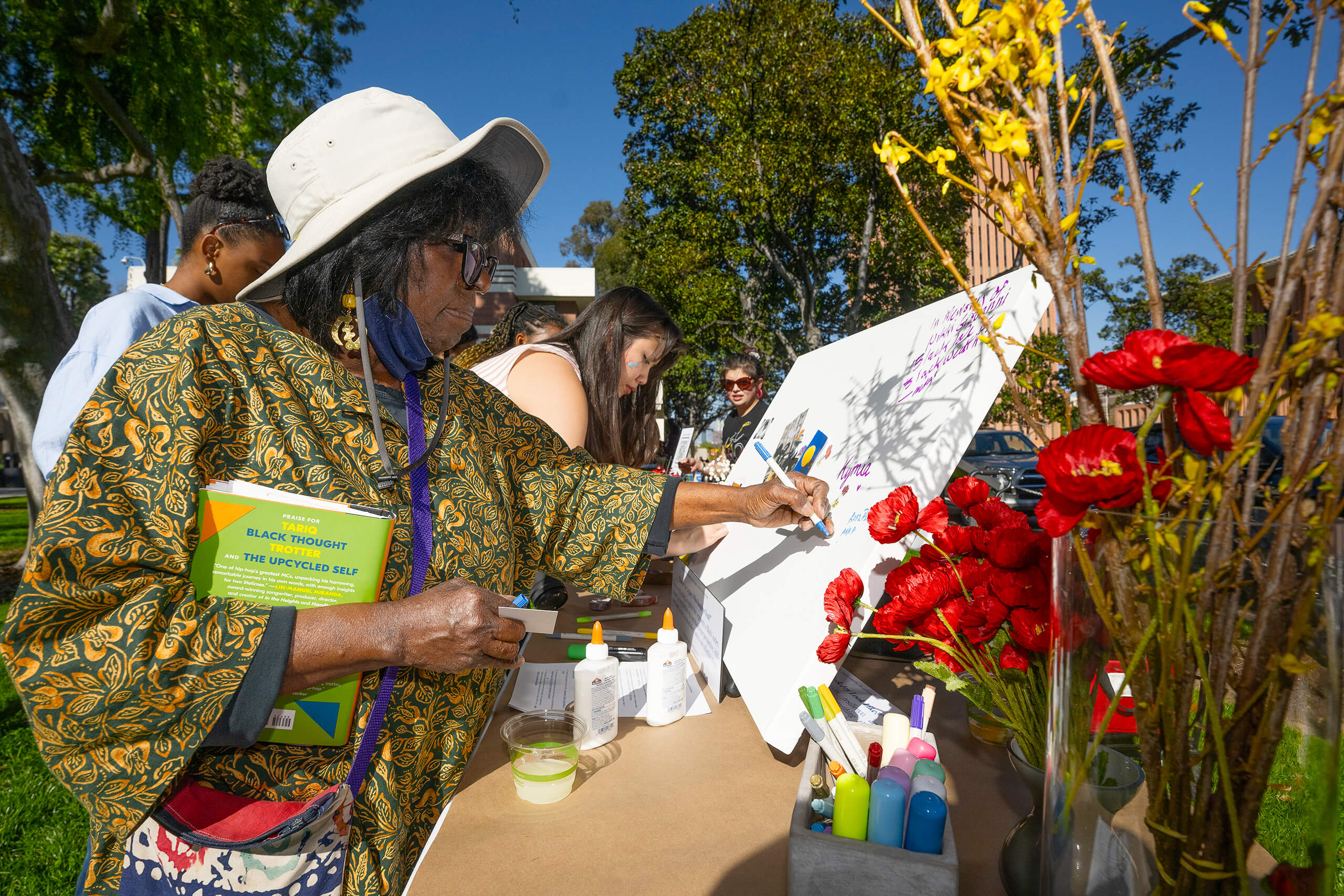 A woman writes on a board at an outdoor event, holding a book and surrounded by art supplies and flowers.